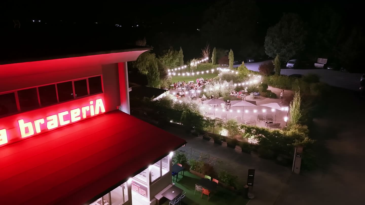 Veduta aerea notturna de La braceriA: l’insegna accesa, il dehors sotto le luminarie e i tavoli pieni